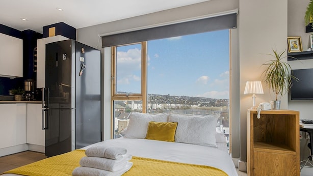 A modern bedroom with a large bed, a view of the city, a kitchen area, and a wooden side table with a lamp. - Luxury Loft Living | Free Parking - Pass the Keys (Brighton)
