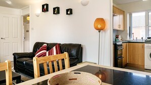 A modern living room with a glass dining table, a black leather sofa, and a woven basket. There is a kitchen visible through an open doorway.