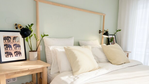 Een netjes opgemaakt slaapkamer met een houten hoofdbord, een nachtkastje met een lamp, een lijst met een afbeelding en twee potplanten.