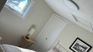 Et soveværelse med en seng, et natbord med lampe, et billede i ramme på væggen og et vindue med udsigt til træer.