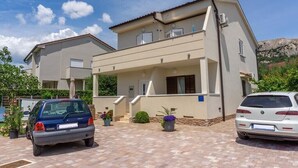 A two-story house with a paved driveway, a car parked in front, and a mountain in the background. - Apartment Adria Nr. 2 with garden and grill (Baska)