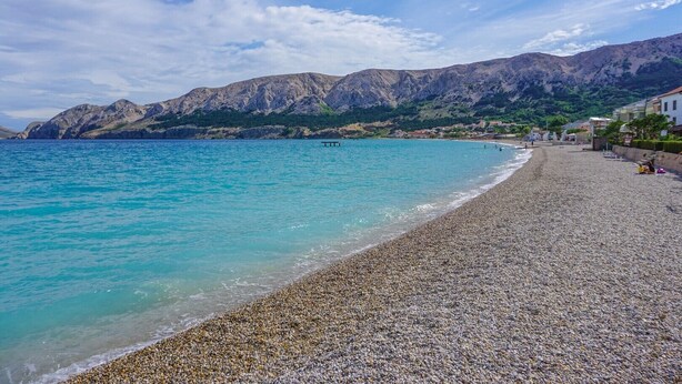 A pebble beach with clear turquoise water, a wooden pier, and mountains in the background. - Apartment Adria Nr. 2 with garden and grill (Baska)