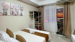 A hotel room with two beds, a wardrobe, a shelf unit, and a view of the city at sunset. - Stunning Mall of Asia View @ Shell Residences (Pasay)