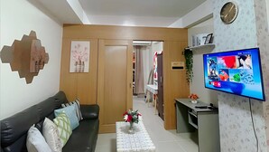 A living room with a black sofa, a coffee table with a vase of flowers, a TV on the wall, and a wooden door leading to another room. - Stunning Mall of Asia View @ Shell Residences (Pasay)