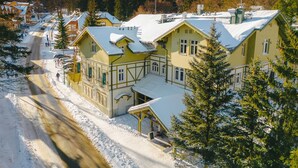 Un grand bâtiment jaune, avec un toit recouvert de neige, entouré d’arbres et avec une montagne en arrière-plan.