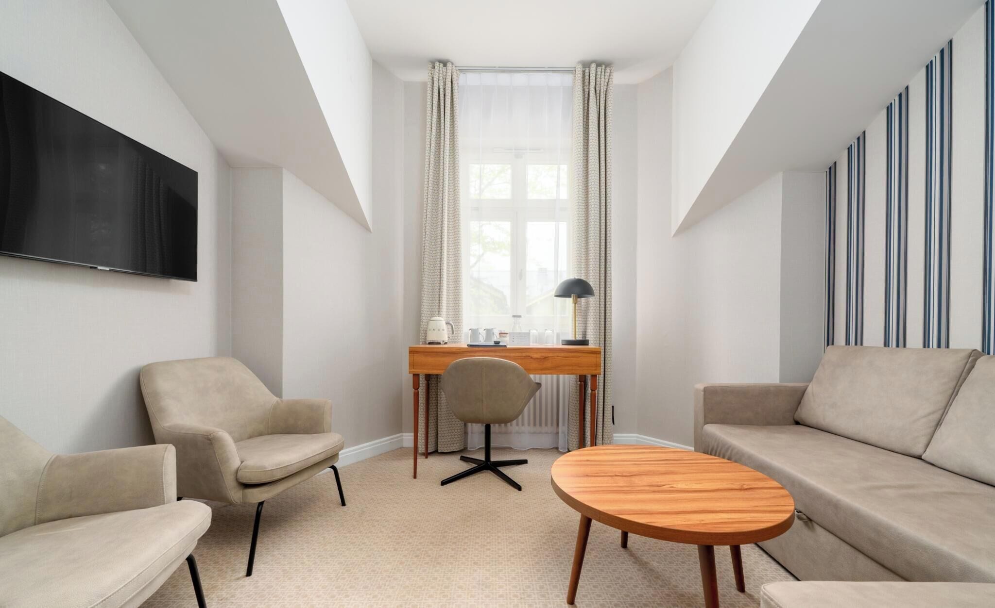 A modern hotel room with a flat-screen TV, a desk with a chair, a round coffee table, and two sofas.