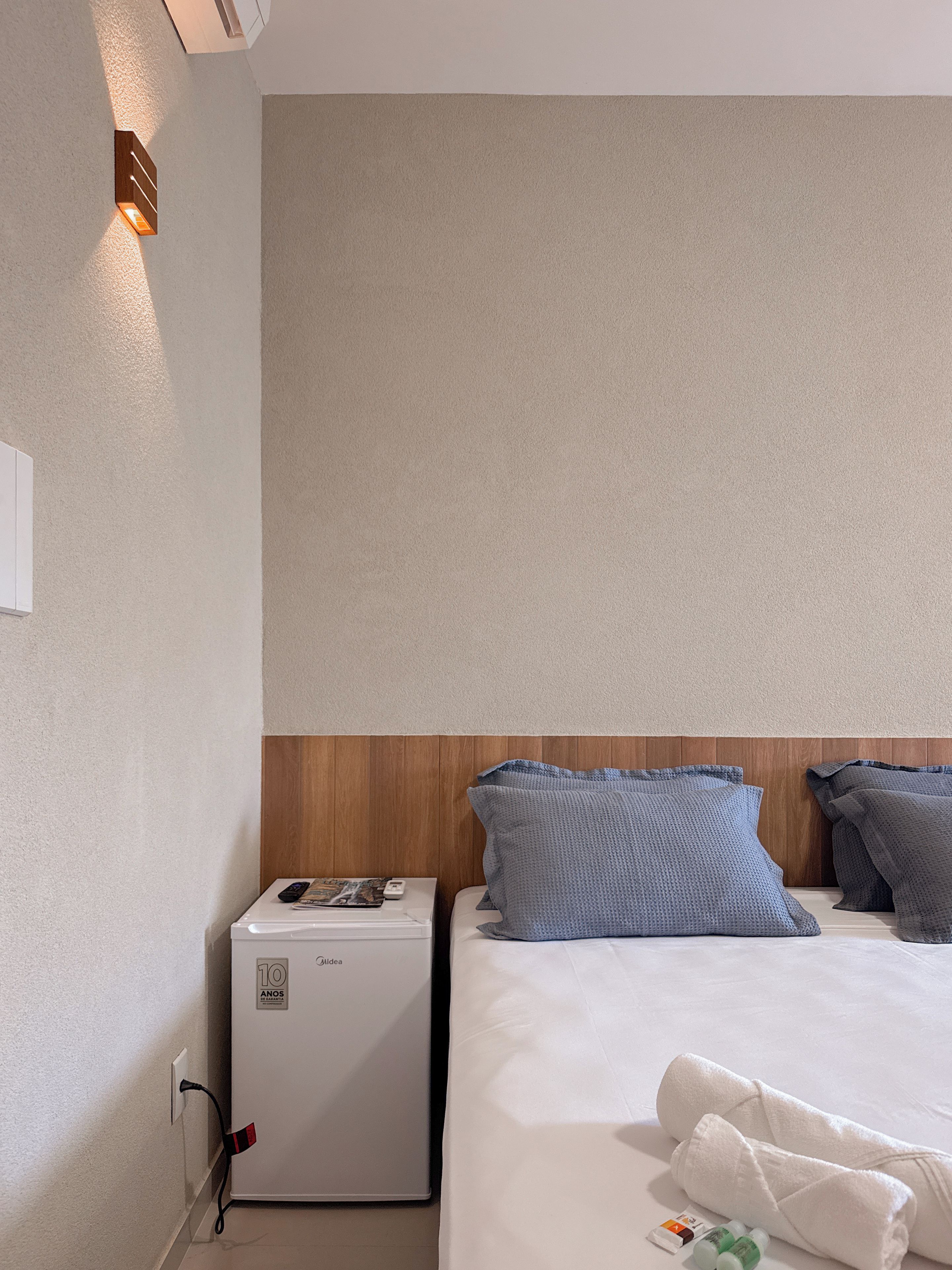 Una habitación de hotel con cama, un calentador pequeño, una mesita de noche y una lámpara fijada en la pared.