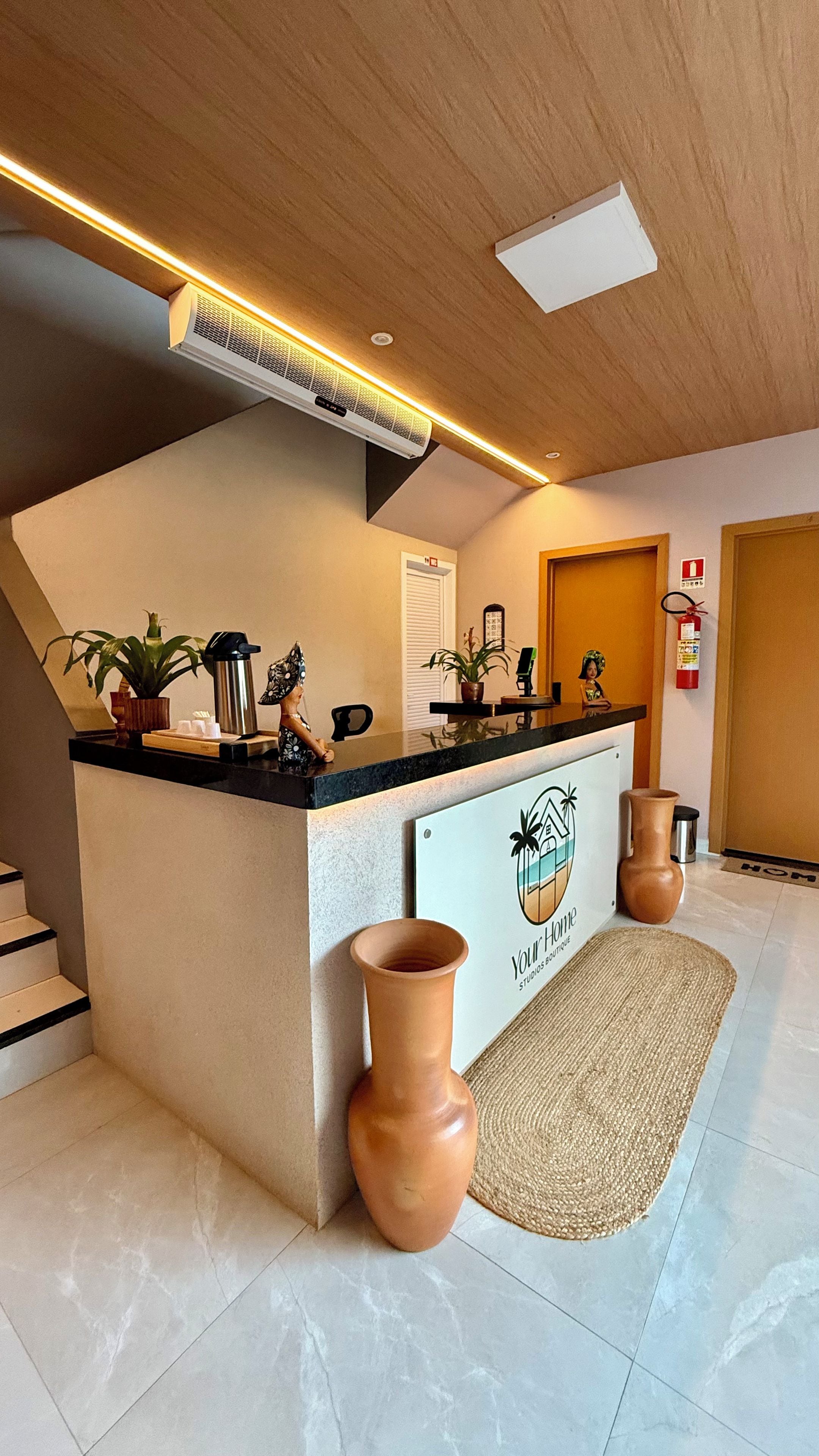 Zona de recepción con mostrador, extintor, planta en maceta, jarra y un cartel que dice 'Your Home Studios Boutique'.