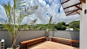A rooftop patio with wooden benches, a large potted plant, and a view of a brick wall and greenery. - Your Home - Studios Boutique Hotel (Sao Luis)