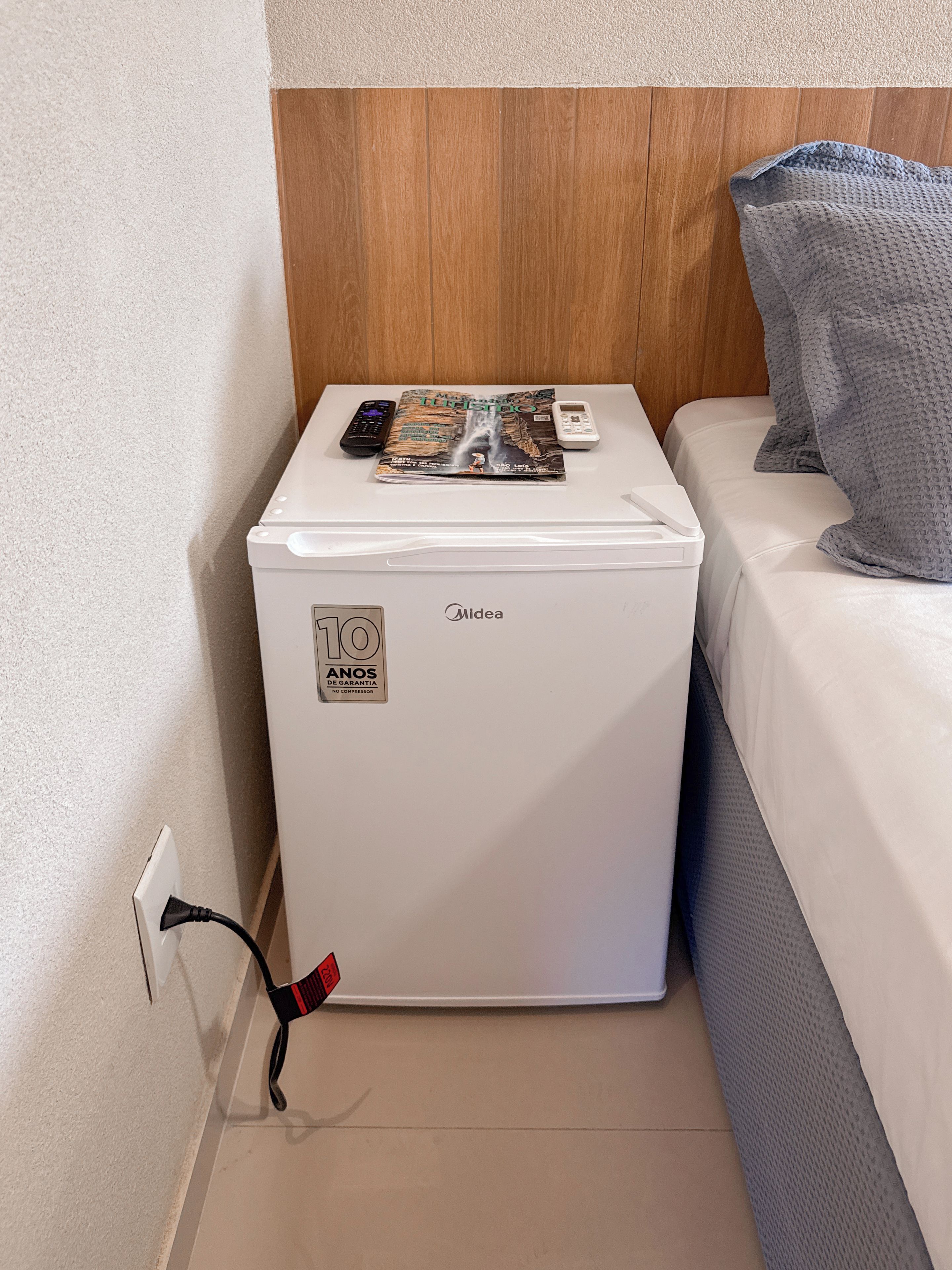 Un appareil blanc et compact, orné d’un autocollant indiquant une garantie de 10 ans, est posé sur une surface blanche, à côté d’un lit recouvert d’une couverture grise.