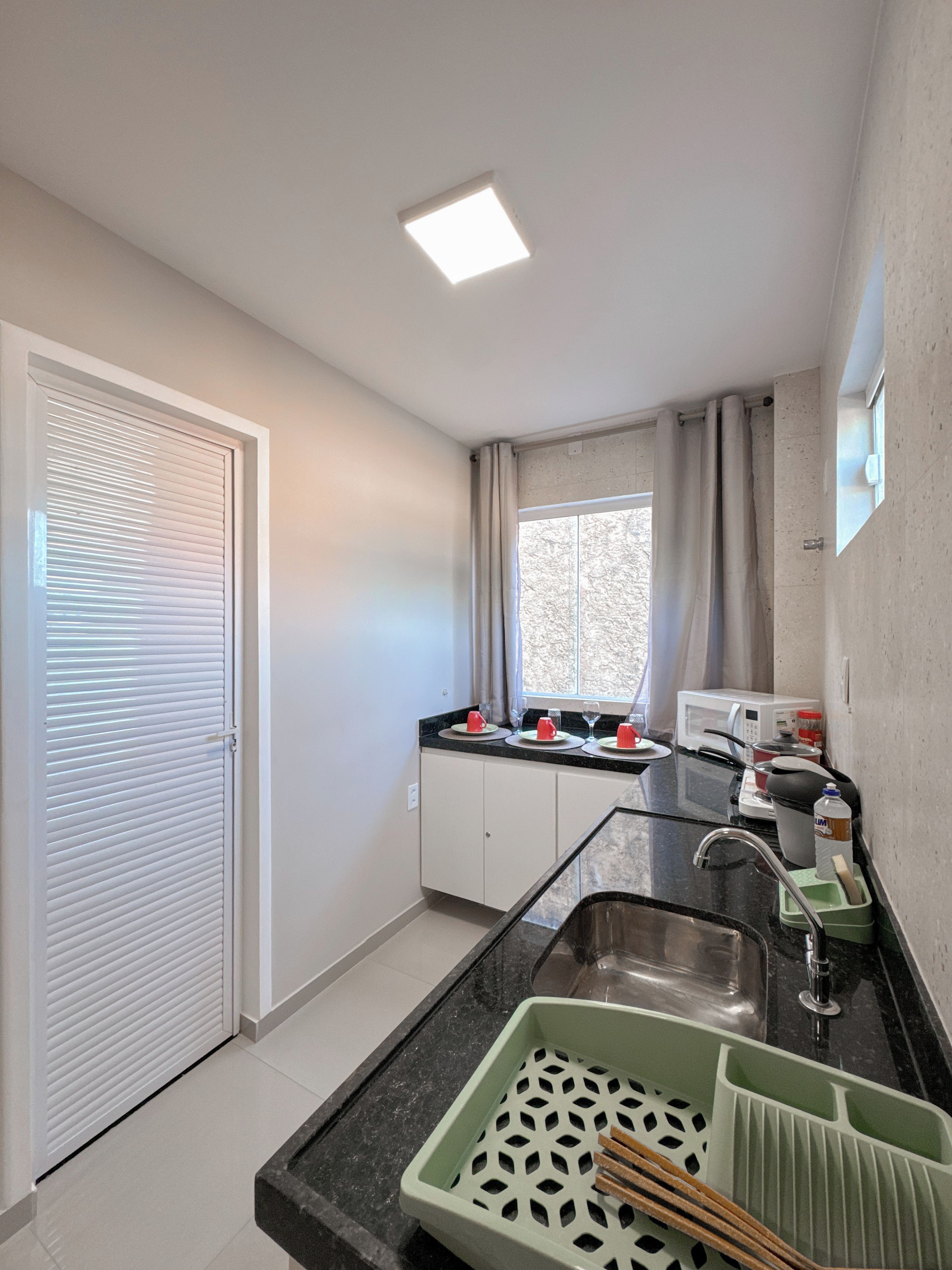 Cozinha compacta com micro-ondas embutido, pia com tapete verde e janela com cortinas.