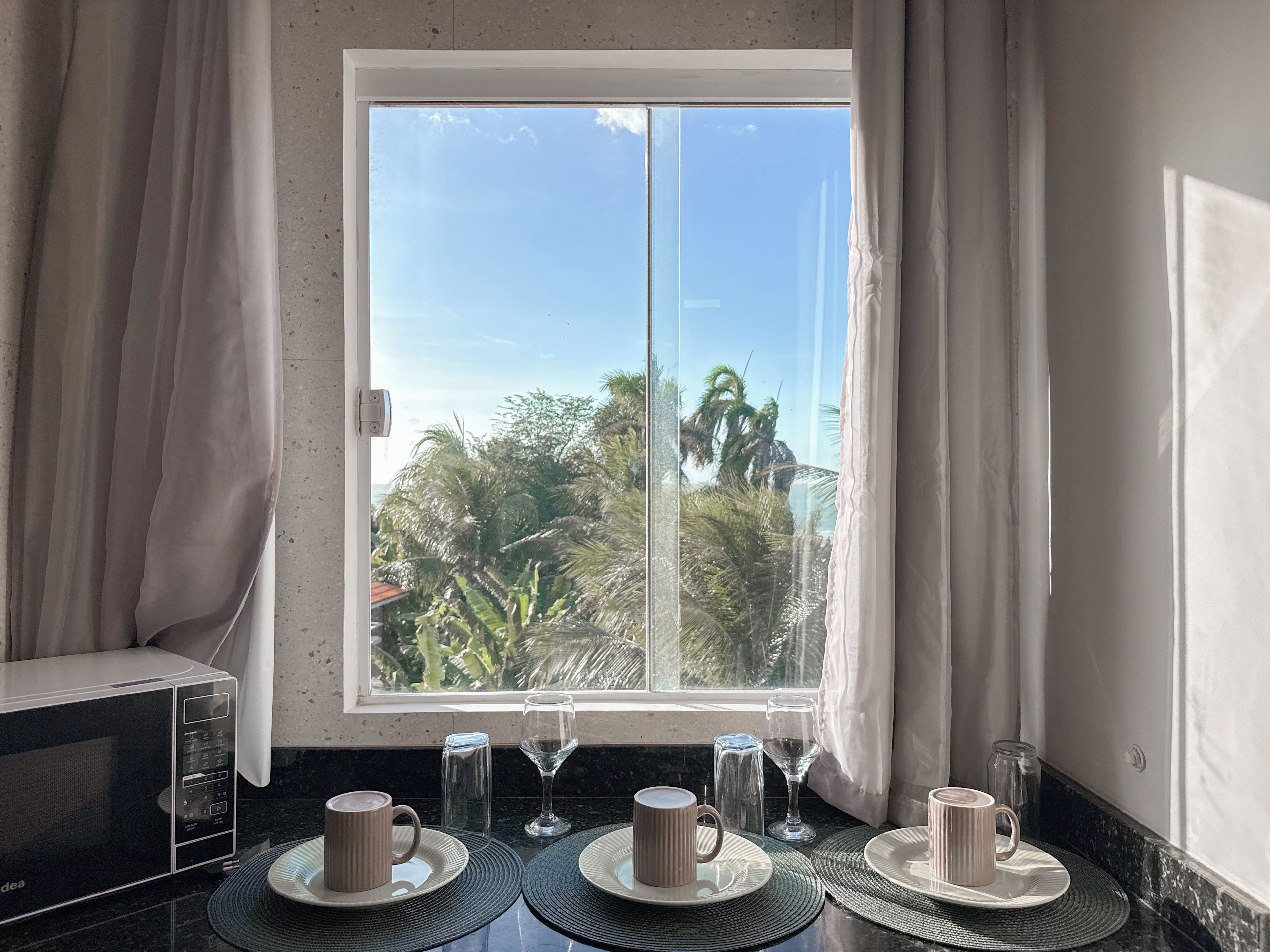 A kitchen with a window overlooking a tropical landscape, a microwave, and a table set with plates, cups, and a glass.