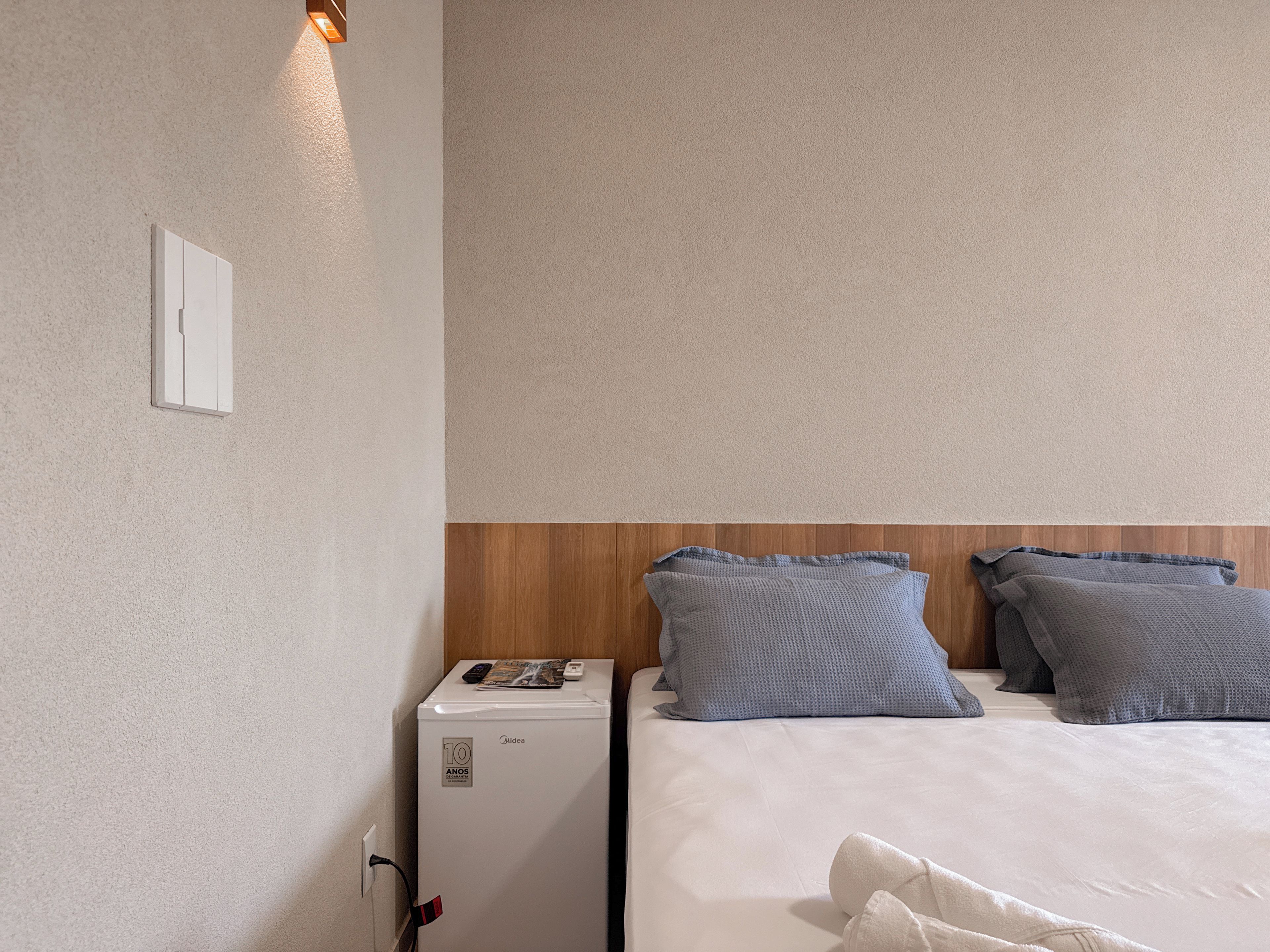 Una habitación de hotel con cama, mesita de noche, un electrodoméstico pequeño blanco y una lámpara fijada a la pared.