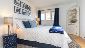 A bedroom with a bed, bedside lamps, a window with curtains, a wall-mounted shelf, and framed pictures.