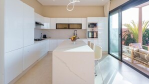 A modern kitchen with white cabinets, a marble island, and a dining area with a glass door leading to a patio. - VILLA YASMINA ( Not overlooked, heated pool) (Marrakech)