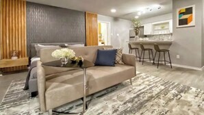 A modern living room with a sofa, a coffee table, and a patterned rug. There is a kitchen area with bar stools and a countertop visible in the background. - Montrose Cheery Studio! Medical Center 09 ★★★★★ (Houston)