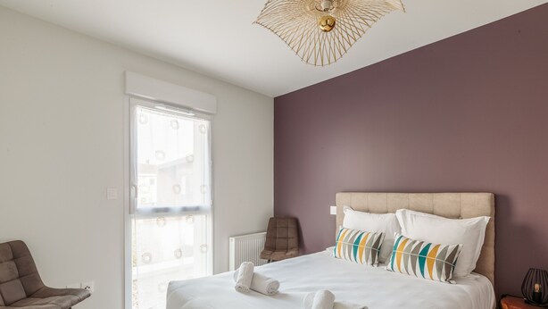 Een slaapkamer met een groot bed, een stoel, een nachtkastje en een raam met jaloezieën.