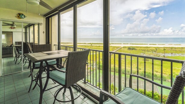 Un balcón con mesa y sillas, con vista a un paisaje costero que incluye el mar y una zona verde.