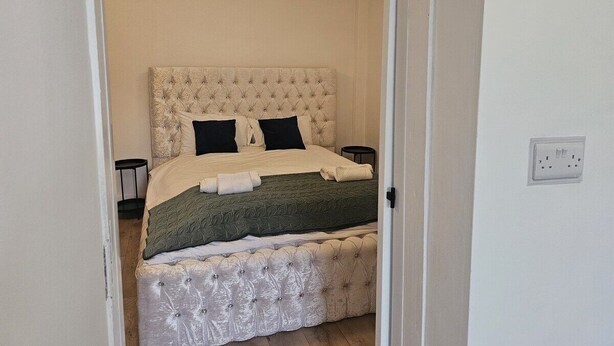 A neatly made bed with a tufted headboard, two bedside tables, and a power outlet on the wall. - Cosy Studio in the Heart of Bridgend (Bridgend County Borough)