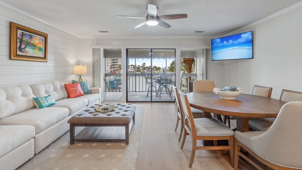 A modern living room with a sofa, ottoman, dining table, and chairs. There is a ceiling fan, a TV mounted on the wall, and a window with a view of buildings and palm trees.