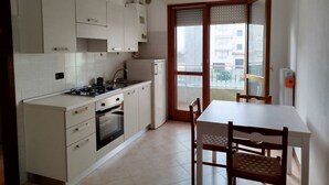 A compact kitchen with white cabinets, a stove, and a refrigerator. There is a dining table with chairs and a view of the outside through a glass door. - Cozy Studio With Pool in Caorle (Caorle)