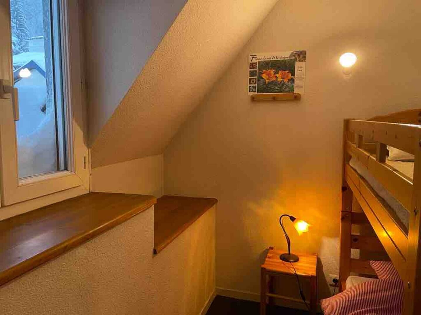 Une petite pièce chaleureuse avec un plafond en pente, un lit superposé en bois, un petit bureau avec une lampe et une fenêtre donnant sur un paysage enneigé.