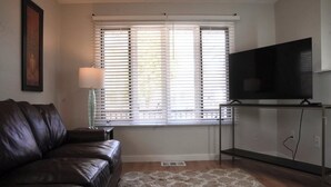 Ein Wohnzimmer mit einer Ledersofa, einem Flachbildfernseher auf einem Schrank, einem Fenster mit Jalousien, einer Stehlampe und einem gemusterten Teppich.
