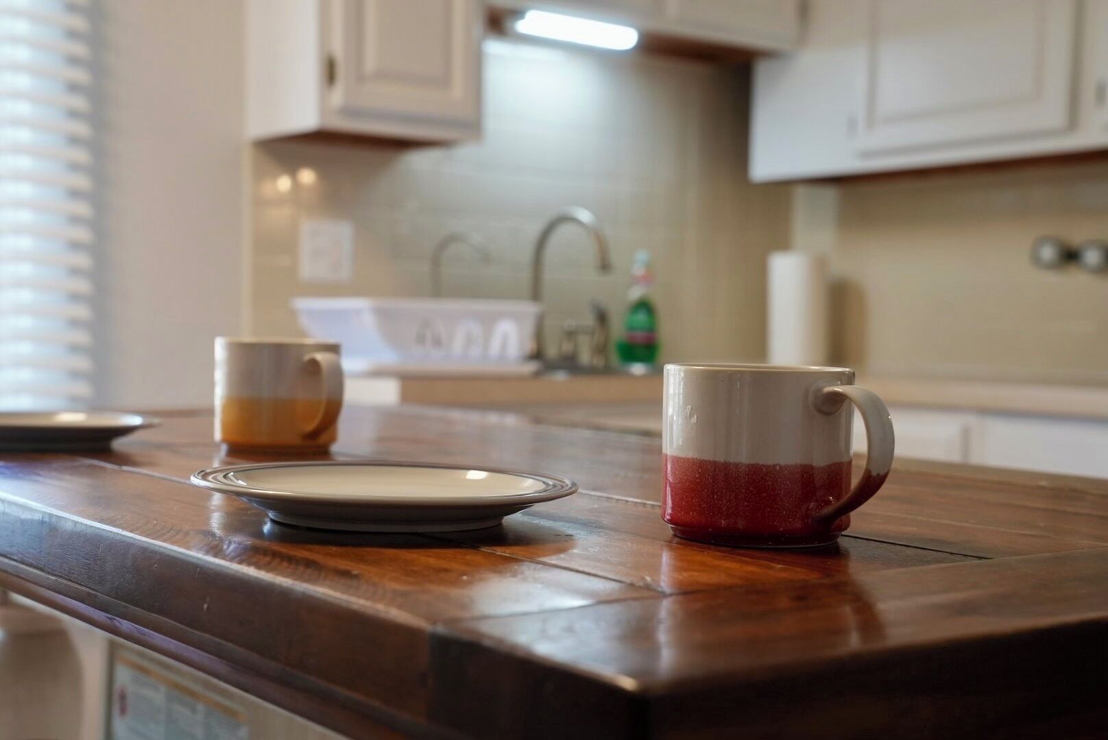 A kitchen counter with a plate and two mugs, a window with blinds, and a sink with a faucet.