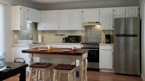 Eine moderne Küche mit weißen Schränken, einem Edelstahl-Kühlschrank, einer Mikrowelle, einem Backofen und einer Kaffeemaschine. Eine Holzeinsel bietet Sitzgelegenheiten, und ein schwarzer Tisch wird von einem Stuhl begleitet.