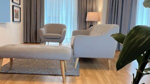 Ein stilvolles Wohnzimmer mit Sofa, Sessel und Hocker, einer großen Pflanze und einem Fenster mit Vorhängen.