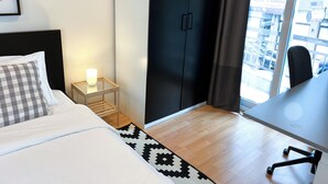 Ein Hotelzimmer mit Bett, Nachttisch mit Lampe, Kleiderschrank, Schreibtisch mit Stuhl und Blick auf die Stadt durch ein Fenster.