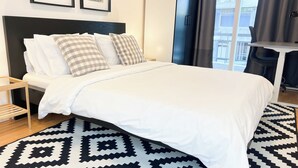 Ein ordentlich bezogenes Bett mit weißer Decke und karierten Kissen, einem schwarz-weißen Teppich und einem Nachttisch aus Holz.