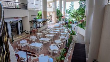 Un patio interior con varias mesas y sillas dispuestas en filas, rodeado de columnas grandes y plantas en macetas.