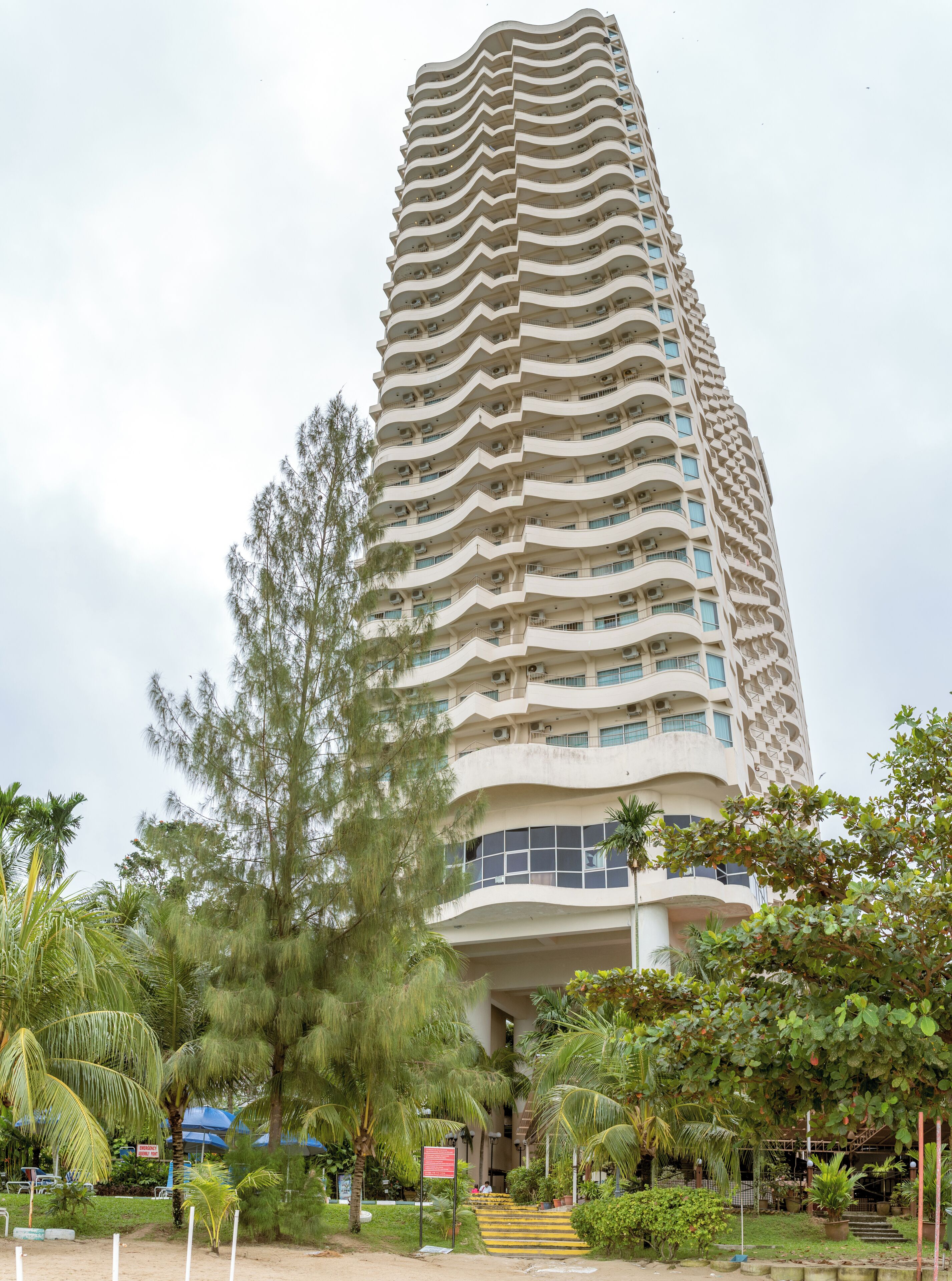 A tall, multi-story building with a unique wavy facade, surrounded by lush greenery and palm trees.