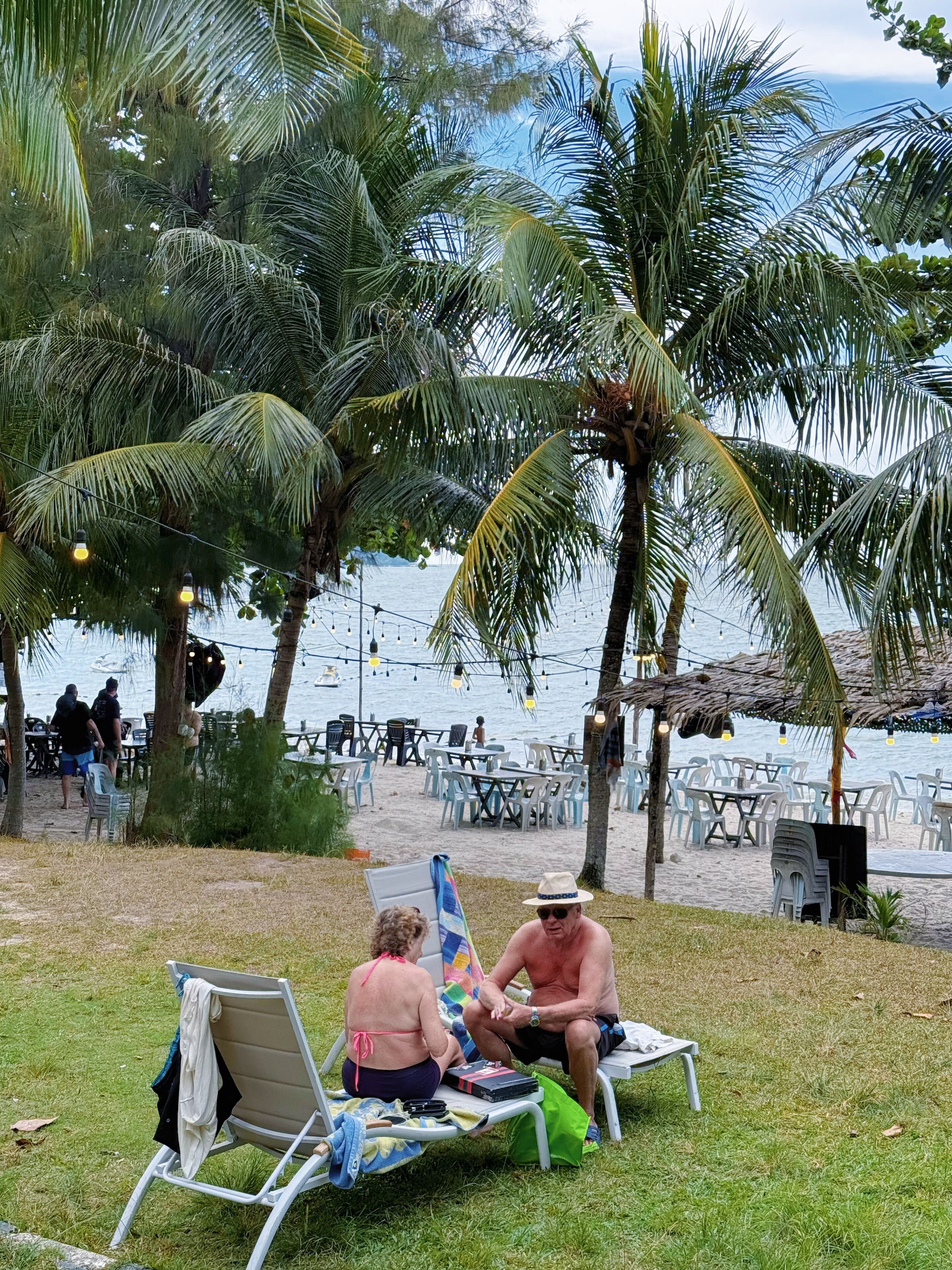 A beachside seating area with lawn chairs and tables, surrounded by palm trees and a view of the sea.
