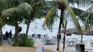 A beachside seating area with lawn chairs and tables, surrounded by palm trees and a view of the sea.