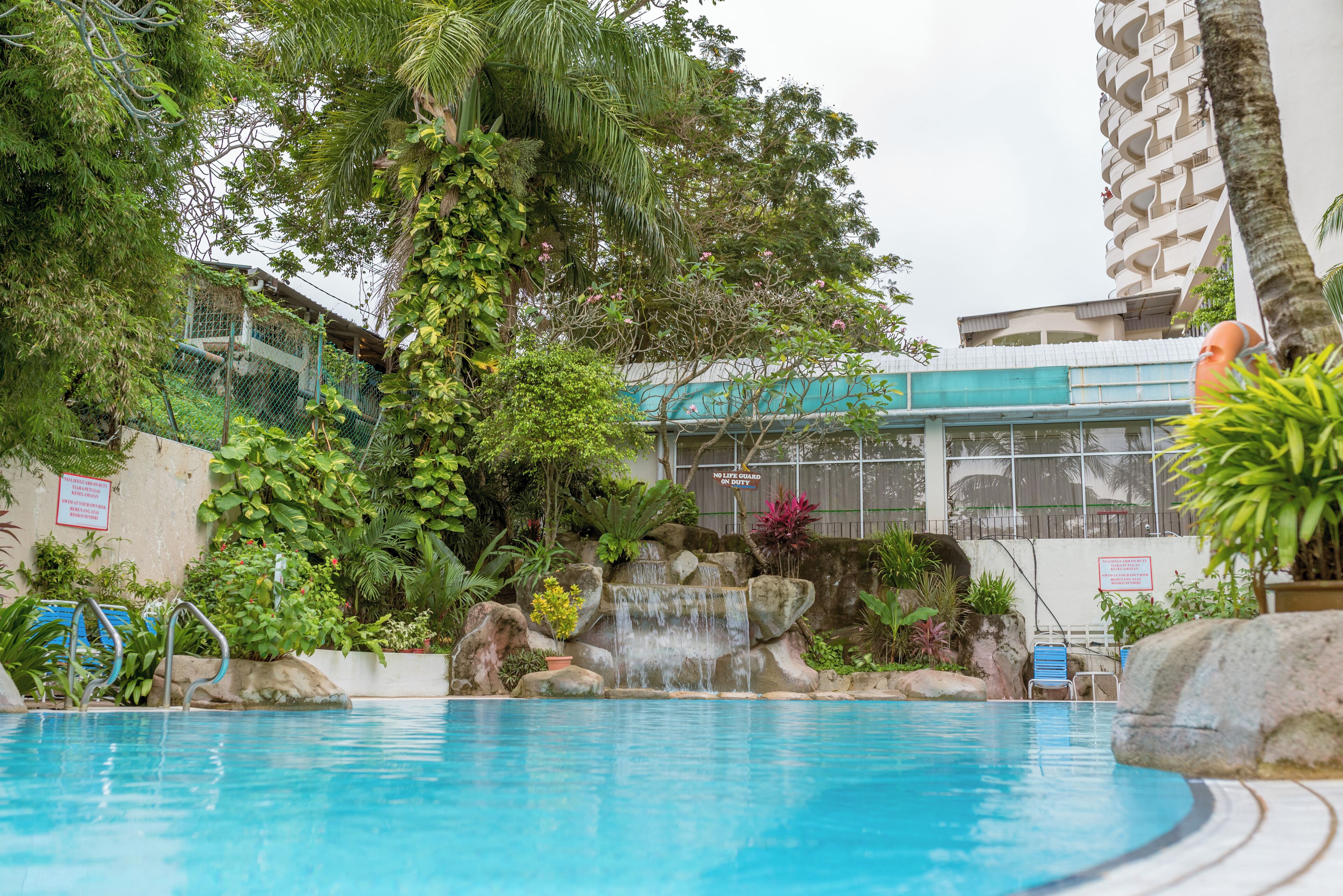 A swimming pool with a waterfall feature, surrounded by lush greenery and a building with balconies.