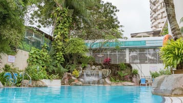 A swimming pool with a waterfall feature, surrounded by lush greenery and a building with balconies.