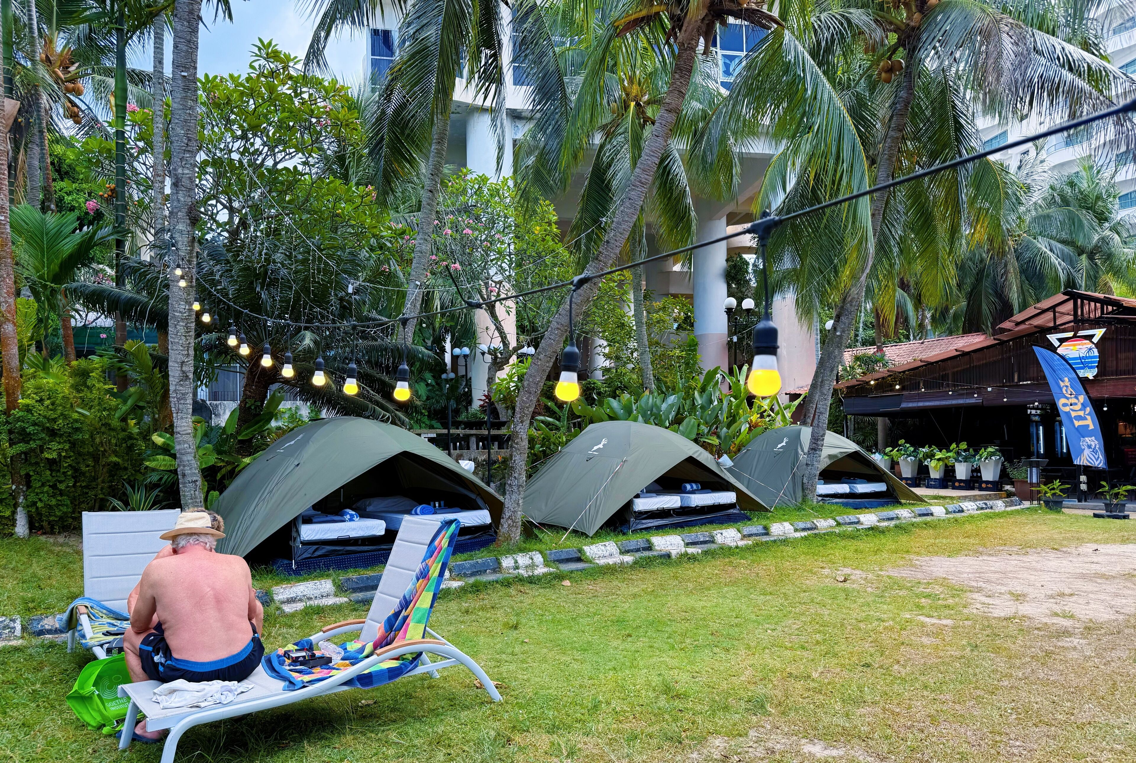 A man sitting on a lounge chair in a tropical garden with several tents and string lights.