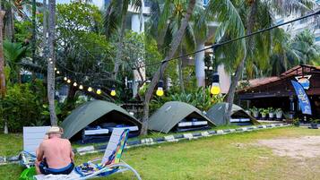A man sitting on a lounge chair in a tropical garden with several tents and string lights.