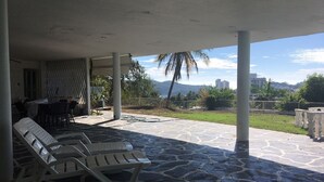 Un'area esterna coperta con pavimento in pietra, poltrone da lounge bianche e vista su una città e montagne sullo sfondo.