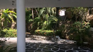 Un cortile con pavimento in pietra, una colonna bianca, rigogliosa vegetazione tropicale e una piccola cascata.