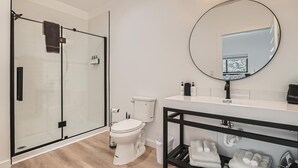 A modern bathroom with a glass-enclosed shower, a toilet, a sink with a mirror, and a shelf with towels. - 2x King Room, A/C, Balcony, Mtn Boutique Motel (Idaho Springs)