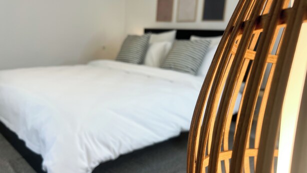 Una cama bien hecha con ropa de cama blanca y almohadas grises, una silla de madera con respaldo trenzado y una pared lisa con tres cuadros.