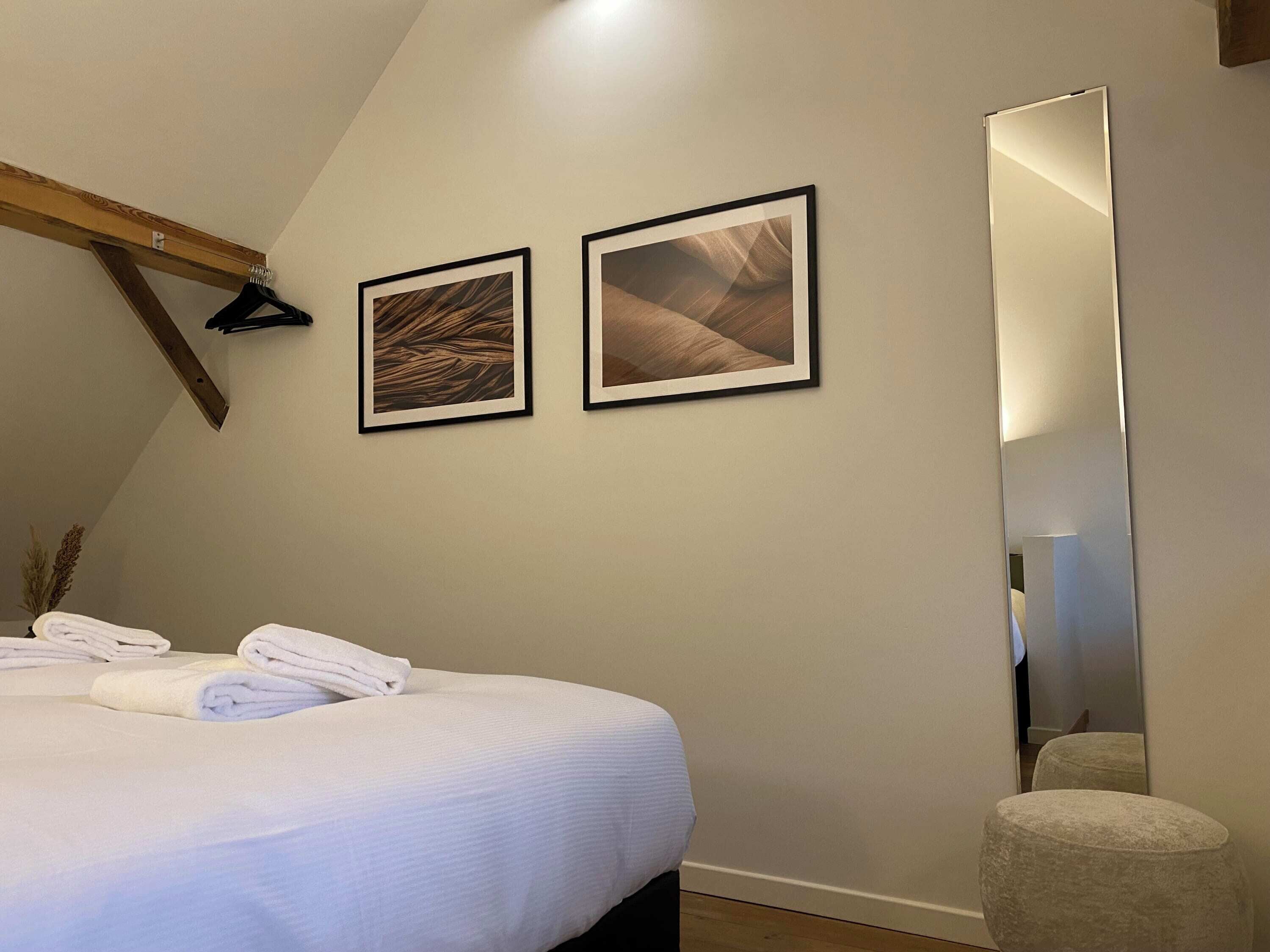 Un dormitorio con una cama, dos cuadros en la pared, un espejo grande y un otoman pequeño y redondo.