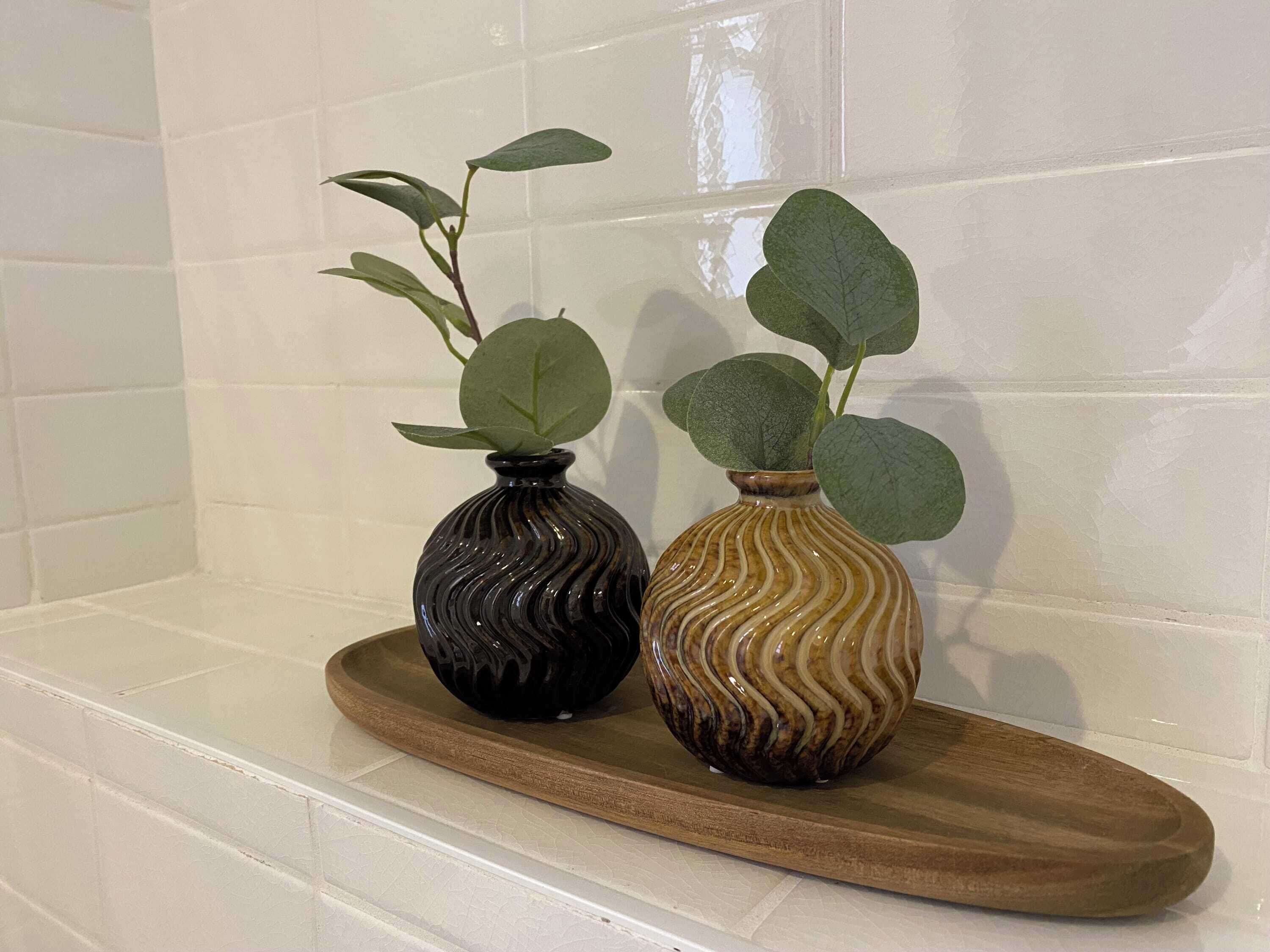 Dos jarrones con plantas sobre una bandeja de madera frente a una pared de azulejos.