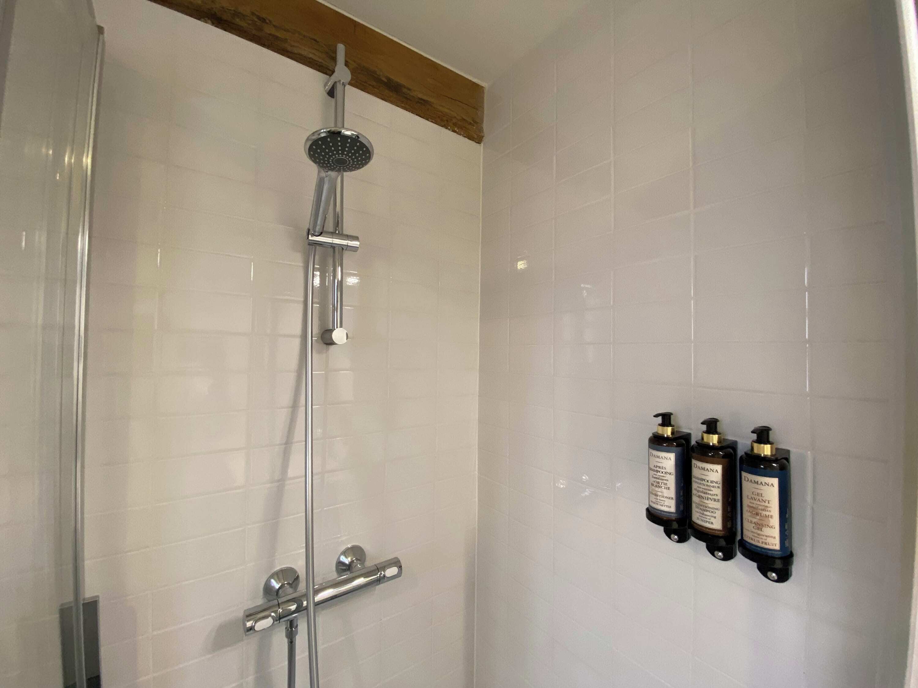 Ducha con cabezal de ducha manual y grifo empotrado en la pared. Hay tres botellas de champú y acondicionador en la pared.