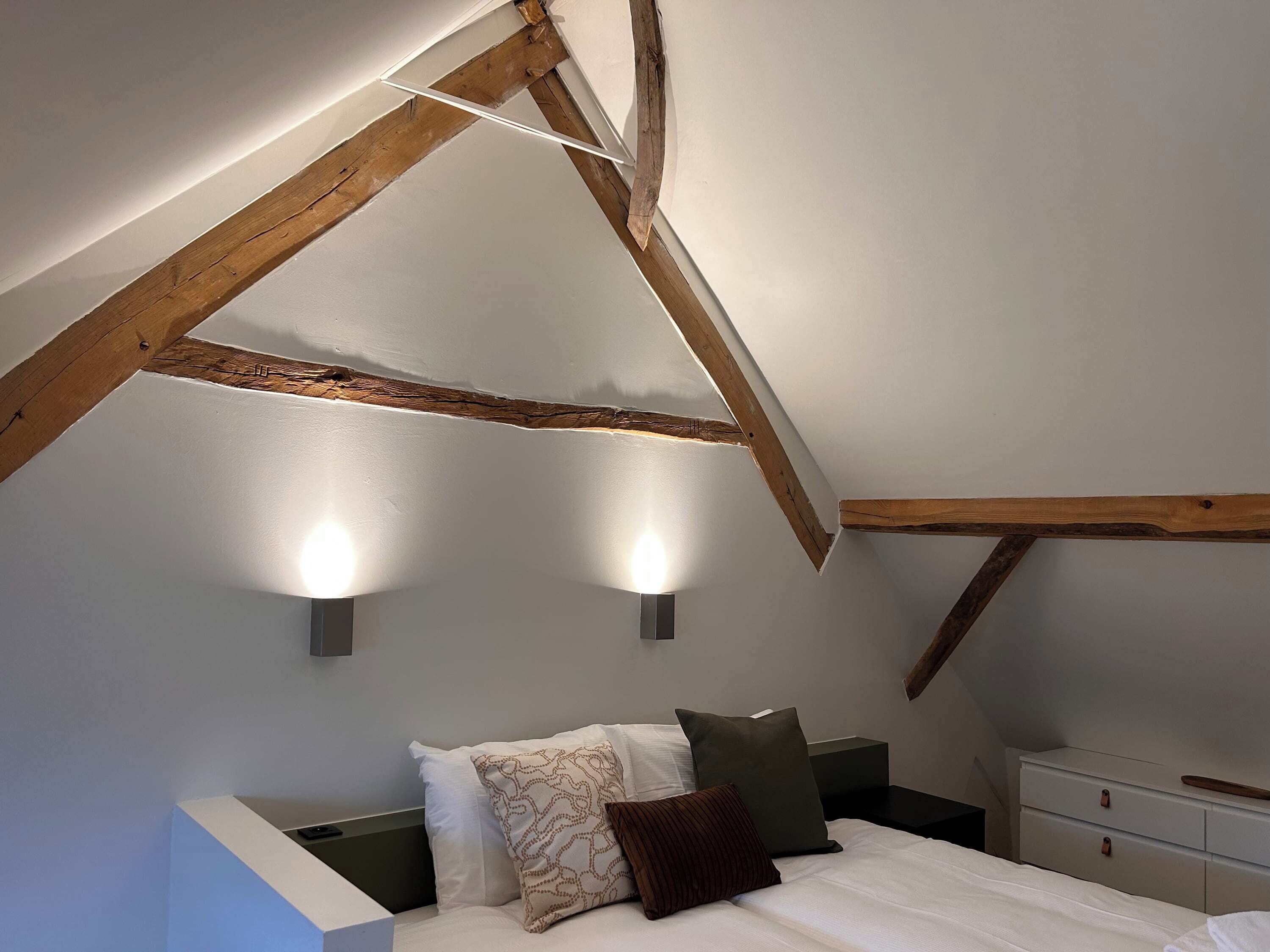 A bedroom with a sloped ceiling, exposed wooden beams, a bed with pillows, a bedside table, and wall-mounted lights.