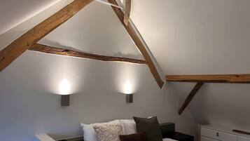 A bedroom with a sloped ceiling, exposed wooden beams, a bed with pillows, a bedside table, and wall-mounted lights.