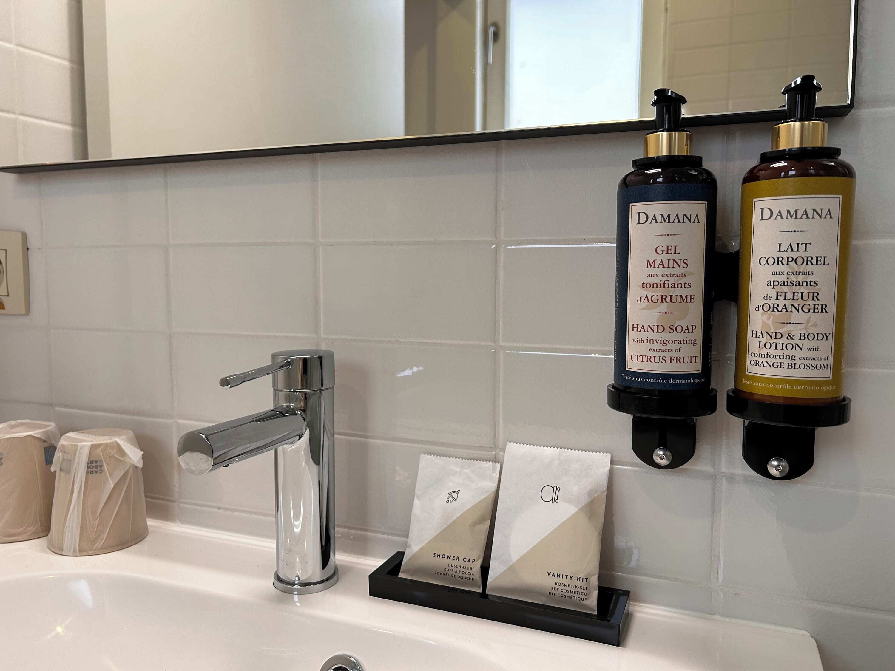 Casa de banho com uma pia branca, torneira cromada e duas embalagens de sabonete e loção para as mãos Damana. Duas pequenas bolsas com a etiqueta “Vanity Kit” estão sobre a pia.